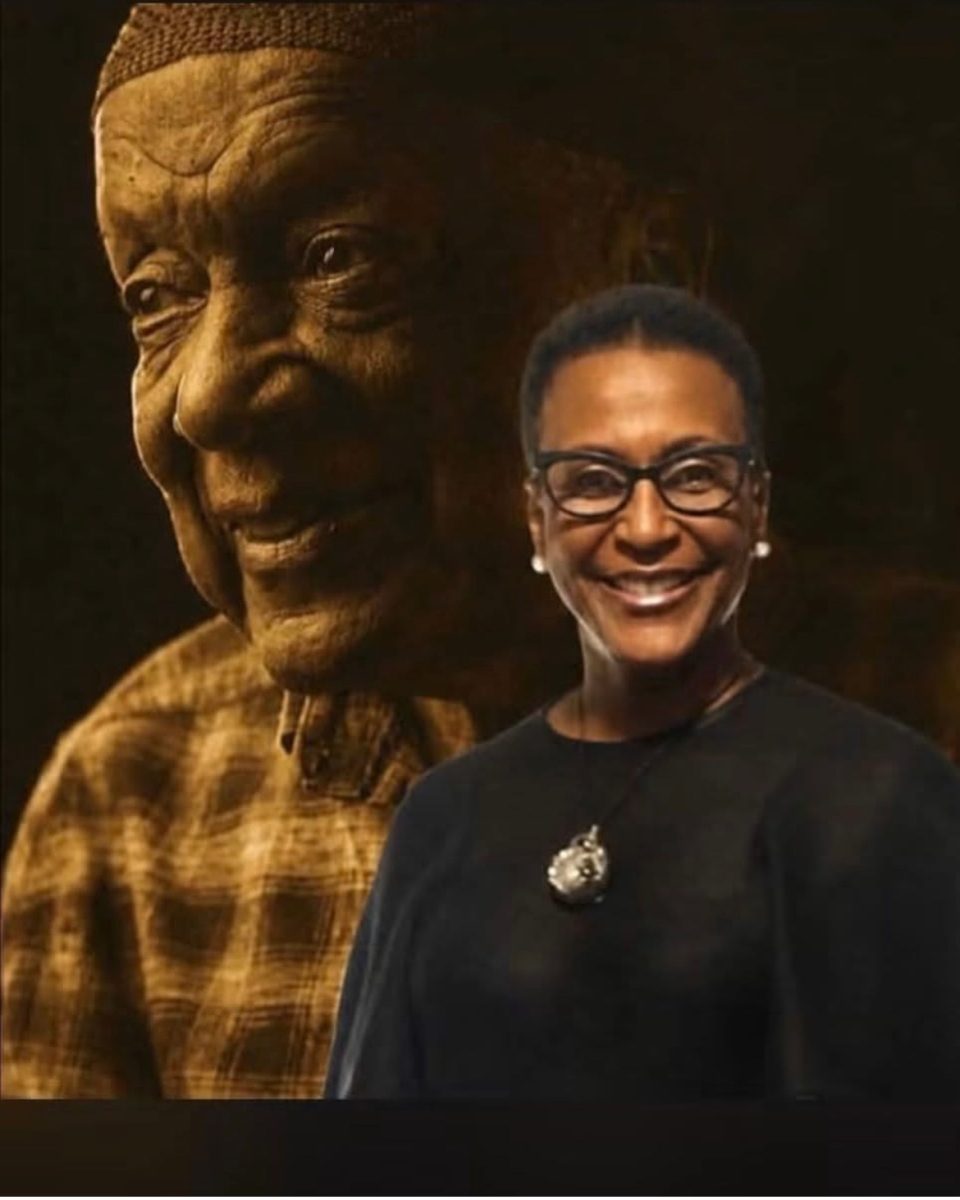 Smiling woman with short hair and glasses in a black top stands in front of a large sepia-toned statue of an elderly man wearing a hat in the background.